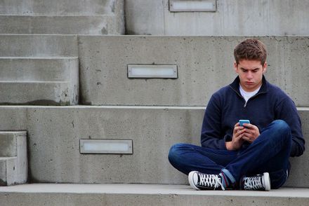 Das Foto zeigt einen jungen Mann, der in einem leeren Sportstadion sitzt und auf sein Handy schaut.