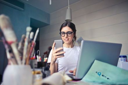 Junge Künstlerin am Arbeitstisch mit Laptop, schaut auf ihr Smartphone, im Vordergrund ein Becher mit Farbpinseln 